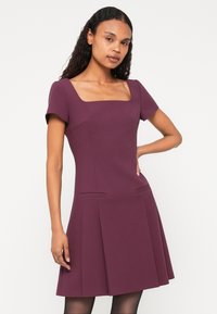 Rochie cu decolteu pătrat, de culoare mov închis. Mâneci scurte, corset ajustat cu buzunare laterale, fustă evazată cu detalii pliate. Textură de material neted.