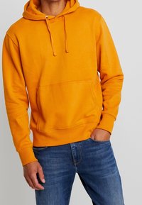 Hombre vestido con una sudadera naranja brillante con bolsillo frontal y jeans azules, con una mano en el bolsillo, frente a un fondo claro y liso.