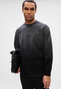 Jovem usando um suéter em degradê cinza a preto, segurando uma bolsa de couro preta com fecho contra um fundo simples.