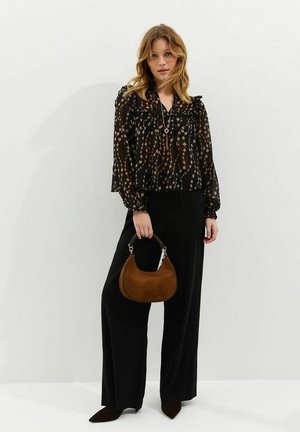 Vrouw in zwarte doorschijnende bloemenblouse en wijde zwarte broek met een bruine suède handtas tegen een witte achtergrond.