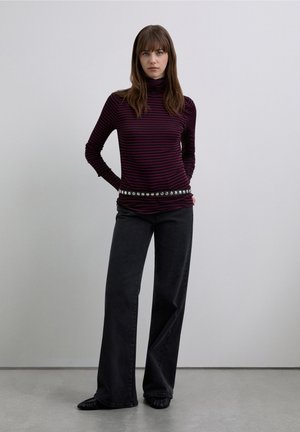 Suéter de cuello alto de manga larga en púrpura oscuro con finas rayas negras, combinado con jeans negros de pierna ancha de talle alto y un cinturón plateado con tachuelas.