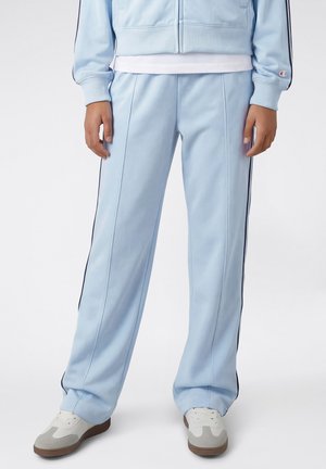 Osoba ubrana w jasnoniebieskie spodnie dresowe z ciemnymi paskami po bokach, białe trampki oraz dopasowaną jasnoniebieską kurtkę na suwak.
