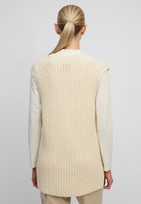 Veste en maille beige clair avec un motif texturé, design sans manches et ourlet arrondi. Portée par-dessus un haut crème à manches longues.