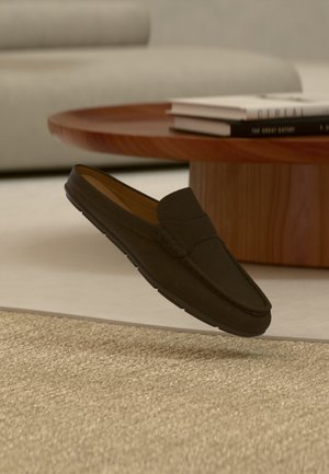 Mørkebrun ruskind slip-on loafer svæver over et beige tekstureret tæppe med et rundt træ sofabord og stabler af bøger i baggrunden.