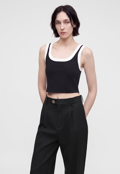 Γυναίκα με κοντά σκούρα μαλλιά φοράει μαύρο cropped τοπ πάνω από λευκό τοπ, συνδυασμένο με μαύρο παντελόνι ψηλής μέσης σε απλό φόντο.