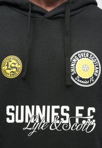 Sweatshirt preta com capuz e tecido texturizado. Apresenta um logótipo circular amarelo com as palavras "Scottish Shiners", texto branco "Sunnies FC" e um desenho de flor.