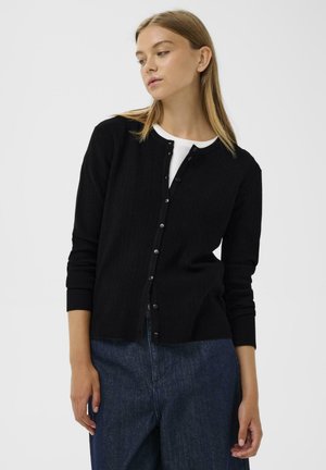 Svart ribbad cardigan med rund hals och knappstängning, med långa ärmar och en figurnära design. Bärs över en vit skjorta.