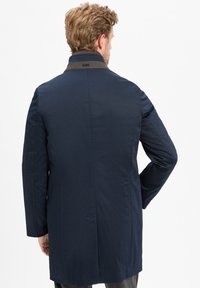 Man in een marineblauwe lange jas met een donkere kraag, gezien van achteren tegen een eenvoudige witte achtergrond.