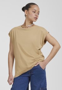 Beige tričko s krátkým rukávem a volným střihem, vyrobené z měkké látky. Mezi výrazné prvky patří ohrnuté rukávy a kulatý výstřih.