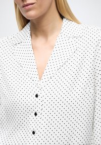 Λευκό πουκάμισο με μαύρες πουά βούλες, διαθέτει γιακά, μπροστινό μέρος με κουμπιά και απαλή υφή. Εμφανή μαύρα κουμπιά στο μπροστινό μέρος.