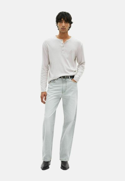 Mann trägt ein weißes langärmliges Henley-Shirt, hellblaue Jeans mit schwarzem Gürtel und schwarze spitze Schuhe, steht mit einer Hand in der Tasche.