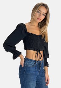 Sort cropped top med firkantet halsudskæring, rynket tekstur, lange pufærmer og bindebånd i livet. Kombineret med blå jeans.