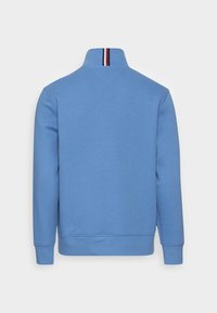 Ljusblå sweatshirt med hög krage. Har röd och vit randig detalj vid halsringningen. Tillverkad av mjukt och glatt tyg.