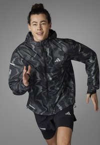 Schwarze und graue gemusterte Laufjacke mit Kapuze, Reißverschluss und reflektierenden Akzenten, kombiniert mit schwarzen Shorts mit einem Logo.