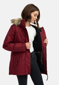 Parka bordeaux con cappuccio in pelliccia sintetica, chiusura a zip e bottoni a pressione, fodera in pile e vita regolabile con cordino. Jeans neri indossati sotto.