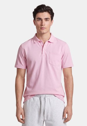 Giovane uomo con una polo rosa chiaro con taschino sul petto e pantaloncini grigio chiaro con coulisse su uno sfondo semplice.