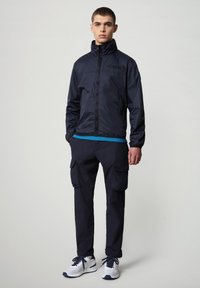 Giacca a vento blu navy con colletto alto, pantaloni cargo coordinati e sneakers bianche. Presenta un design semplice e tasche funzionali.