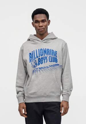 Jeune homme portant un sweat à capuche gris avec l'inscription « Billionaire Boys Club » et un motif d’astronaute bleu, debout devant un fond blanc uni.