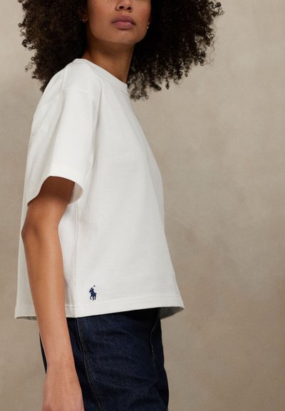 Kobieta z kręconymi włosami w luźnej białej koszuli z krótkim rękawem, z małym granatowym logo polo przy krawędzi oraz w ciemnoniebieskich dżinsach.