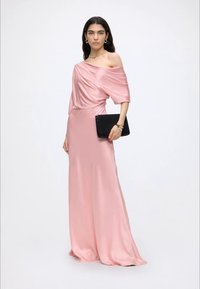 Kvinde iført en lang lyserød satin off-shoulder kjole, der holder en sort clutch taske mod en ren hvid baggrund.