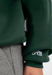 Manche de sweat-shirt vert foncé avec broderie blanche "P.O.B." et étiquette noire portant la mention "PROHIBITED", assortie à un pantalon en laine gris.