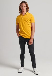 Gul polo-shirt med krave, korte ærmer og en diskret logo, parret med tætsiddende sorte jeans og hvide sneakers. Ensfarvet baggrund.