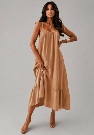 Femme aux longs cheveux foncés portant une robe midi ample beige sans manches et des sandales à talons hauts transparentes, posant avec une main sur la hanche.