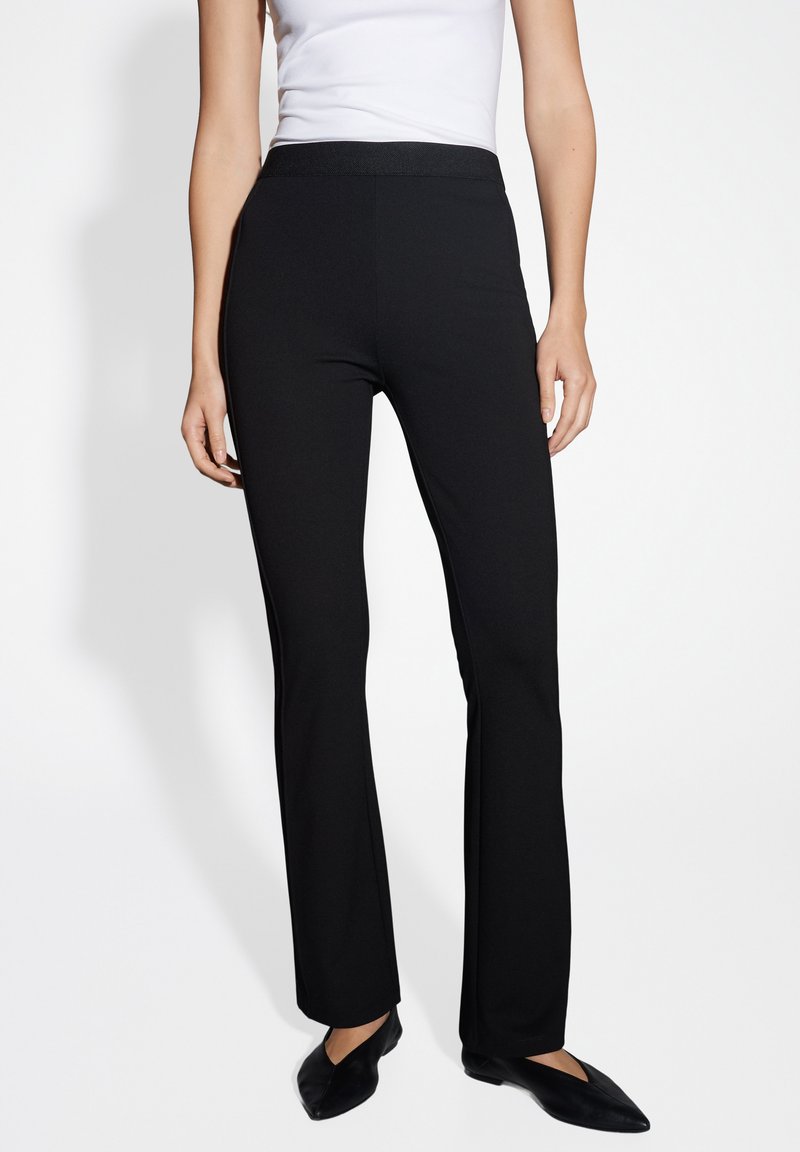 Schwarze Schlaghosen aus glattem Stoff, mit einem elastischen Bund und einem schlichten Design. Kombiniert mit schwarzen spitzen Ballerinas für einen Kontrast.