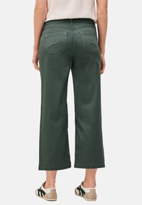 Pantalones de algodón de pernera ancha en verde oliva, con bolsillos traseros, una cintura de media altura y una textura suave. Combinados con zapatillas crema y verde oscuro.