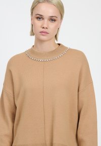 Maglione beige con colletto alto, caratterizzato da una fila di decorazioni in cristallo trasparente attorno al collo. Tessuto morbido e vestibilità ampia.