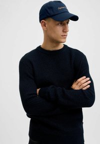 Navy knitted sweater with a textured pattern and ribbed cuffs, paired with a dark blue cap featuring embroidered text on the front.