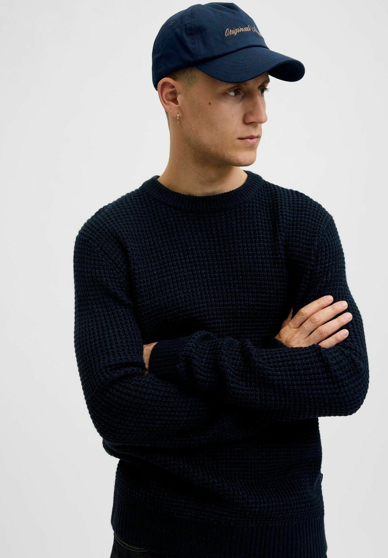 Navy knitted sweater with a textured pattern and ribbed cuffs, paired with a dark blue cap featuring embroidered text on the front.