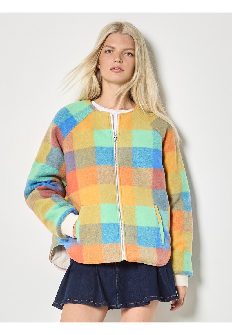 Femme blonde portant une veste zippée à carreaux colorés sur un haut blanc et une jupe bleu foncé, se tenant devant un fond blanc uni.