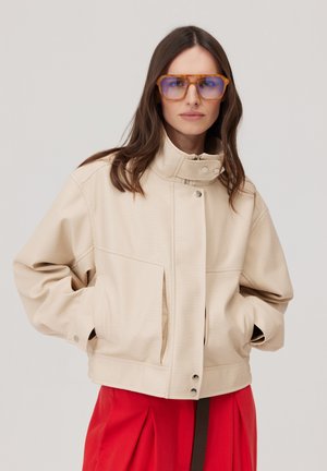 Kobieta ubrana w za dużą beżową kurtkę i duże niebieskie okulary z bursztynową oprawą, stoi z rękami w kieszeniach kurtki.