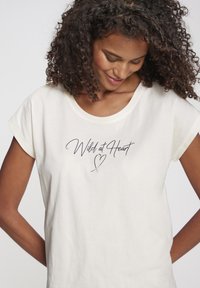 Cremfarbener T-Shirt mit kurzen Ärmeln, rundem Halsausschnitt, mit dem Text "Wild at Heart" in schwarzer Schrift und einem Herz-Graphic.