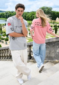 Homme en t-shirt graphique gris et pantalon blanc, debout les bras croisés, femme en t-shirt graphique rose et jean marchant sur des marches en pierre dans un parc.