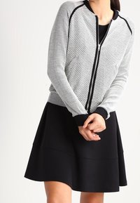 Svart och vit bomberjacka med geometrisk stickad textur, dragkedjestängning, svarta ribbade muddars, bärs över en svart passform och utställd klänning.