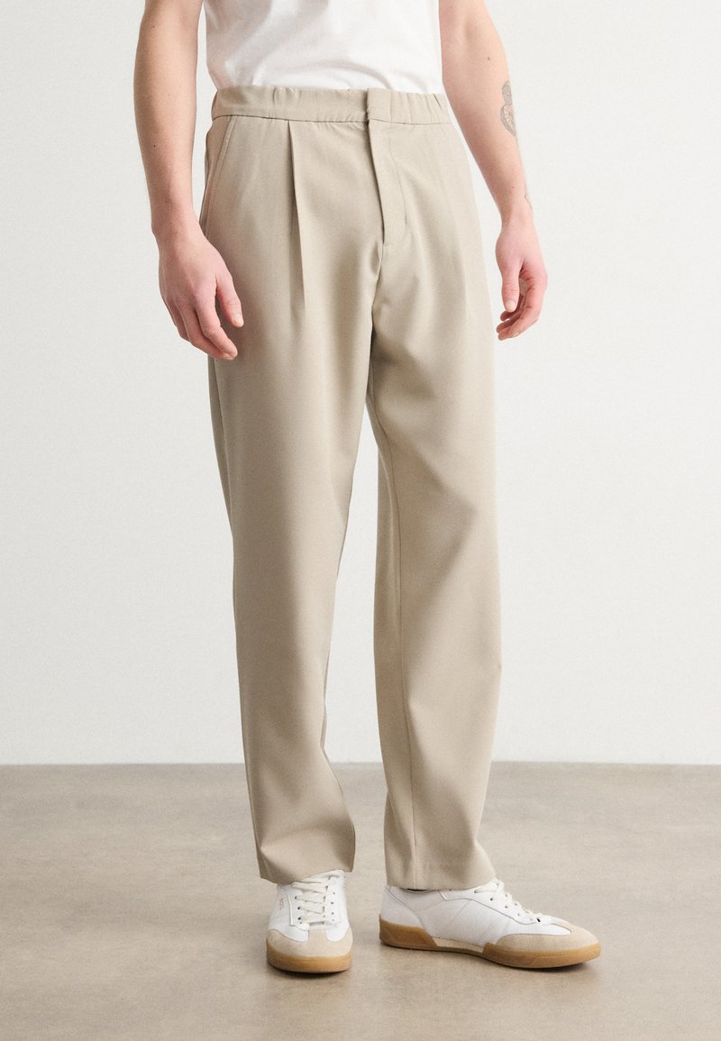 Beige, plisserte bukser med elastisk midje, laget av et glatt stoff. Brukt med hvite joggesko med gummisåle.