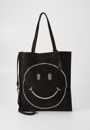 Black suede tote bag with silver studded smiley face design and dual shoulder straps against plain background.