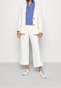 Blazer blanc, sweat à capuche zippé bleu et pantalon blanc à jambes larges. Assorti à des baskets légères avec des accents gris et verts.