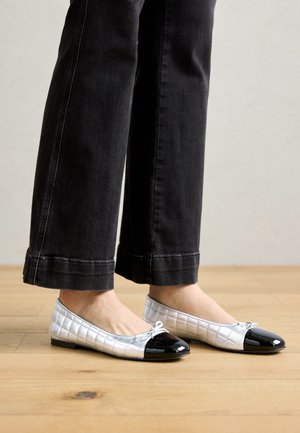 Fødder iført sølvfarvede quiltede balletflats med sorte lak-tåhætter og små sløjfer, kombineret med sorte cropped bukser, stående på trægulv.
