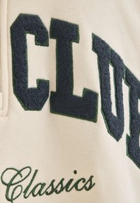 Tessuto crema con lettere in peluche grigio scuro che formano la parola "CLUB" e accenti verdi. La parola "Classics" è ricamata in un font corsivo verde.