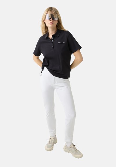 Černé polo tričko s límečkem a knoflíkovou léga, zdobené bílým vyšívaným textem. V kombinaci s bílými slim-fit kalhotami a masivními béžovými teniskami.