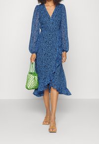 Modré květinové šaty s volným střihem a dlouhými nabíranými rukávy, s volánkovým lemováním, kombinované se zelenou tkanou kabelkou a béžovými sandály na pásky.