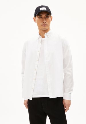 Witte overhemd met lange mouwen, gedragen over een wit T-shirt. Zwarte pet met een rechthoekig logo. Effen zwarte broek.