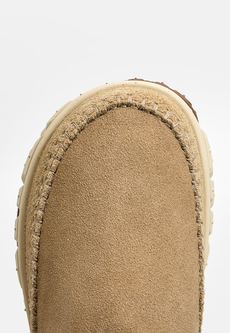 Zapatilla de ante marrón con punta redondeada, suela de goma color crema, costuras visibles a lo largo del borde y una textura suave.