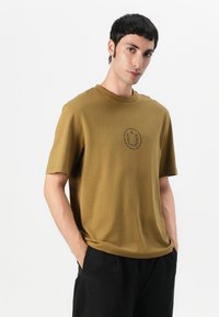Olive green short-sleeve T-shirt made of cotton. Features a round neckline and a smiley face graphic in black on the center.