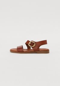 Brown leather sandal with two wide straps and a round gold buckle. Flat sole with a smooth texture. Open-toe design.