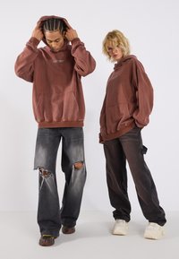 Hnedé oversized mikiny s kapucňou a prednými vreckami, nosené s tmavými džínsami s roztrhnutými kolenami. Uvoľnený, pohodlný strih s viditeľnými šnúrkami.