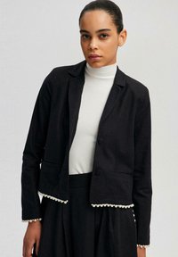 Černý zkrácený blazer s perlami, přizp�ůsobený design, zapínání na jeden knoflík a tenké klopy, spárovaný s bílým topem s vysokým límcem.